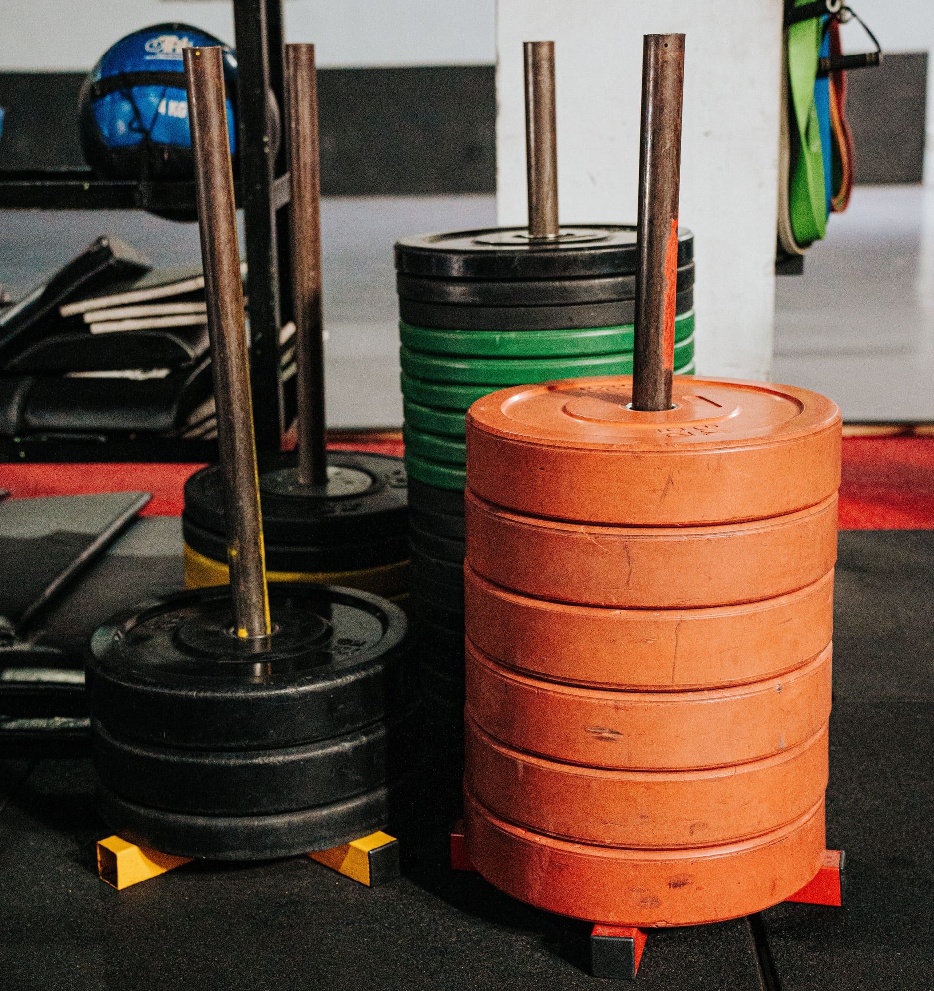 Plate posts on a gym floor stacked with rubber bumper plates: six thick orange plates in front, black and green bumpers behind.