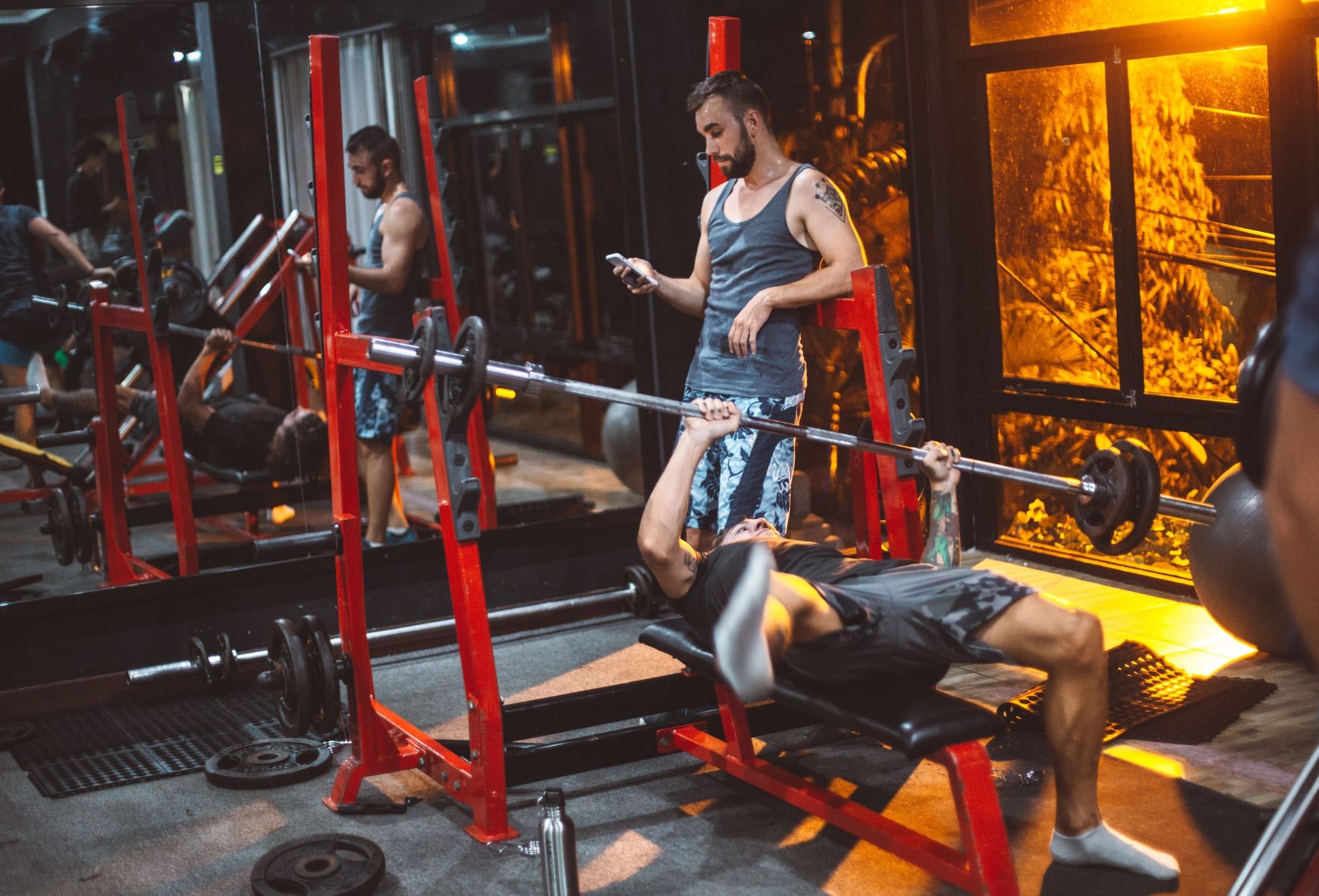 A man bench pressing on a red bench with uprights built into the frame while another man stands at the head of the bench looking at his phone. Plates and a loaded bar lie on the floor around them in a dim commercial gym.