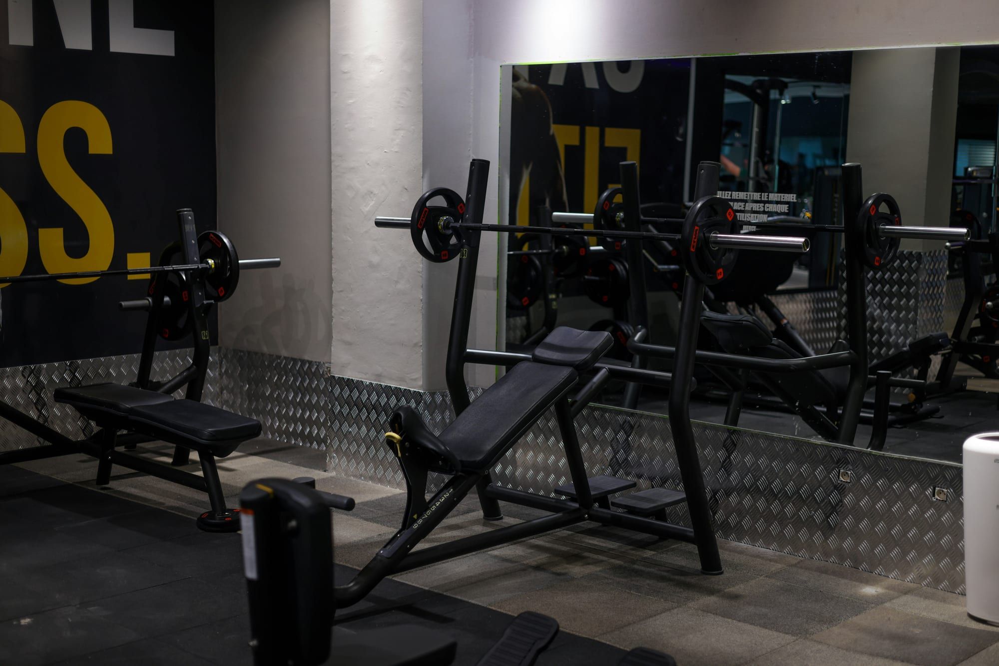 A flat and an incline bench press station in a dimly lit commercial gym, each with uprights built into the frame and a loaded barbell resting on the hooks.