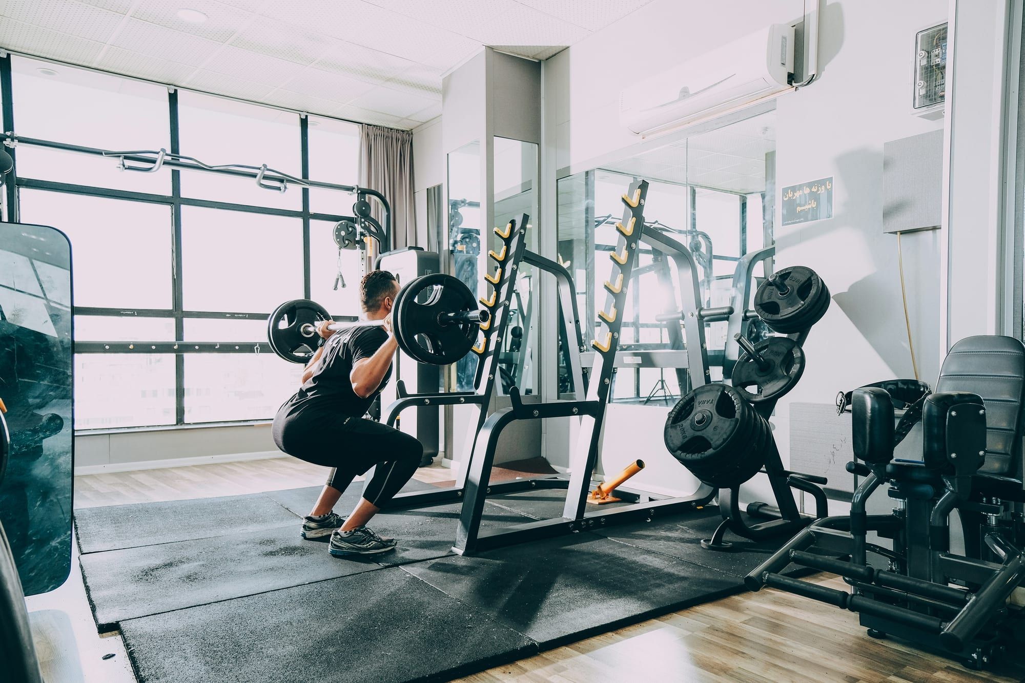 A man squatting with a loaded barbell in a bright commercial gym, beside a squat stand with a column of yellow hooks fixed up each upright.